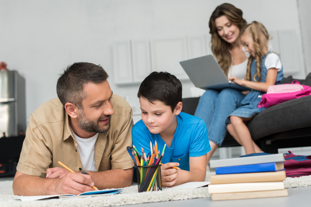 Selektivní fokus člověka pomáhá syn s domácími úkoly, zatímco matka a dcera pomocí notebooku na pohovce společně doma - Fotografie, Obrázek