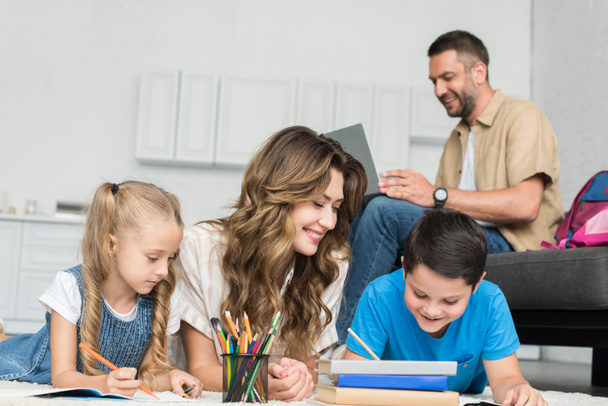mère souriante aider les enfants avec des devoirs tandis que l'homme utilisant un ordinateur portable sur le canapé à la maison
 - Photo, image