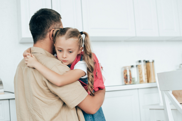 ο πατέρας και θλιβερή μικρή κόρη με σακίδιο που αγκαλιάζονται μεταξύ τους στην κουζίνα στο σπίτι, πίσω στο σχολείο έννοια - Φωτογραφία, εικόνα