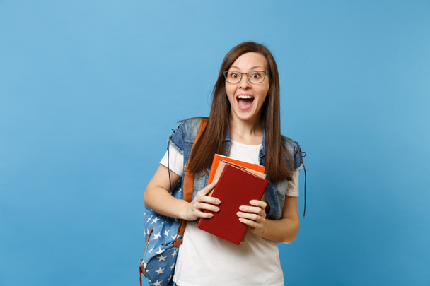 Portrait of young surprised amazed happy woman student in glasses with opened mouth with backpack holding school books isolated on blue background. Education in high school university college concept - Photo, image