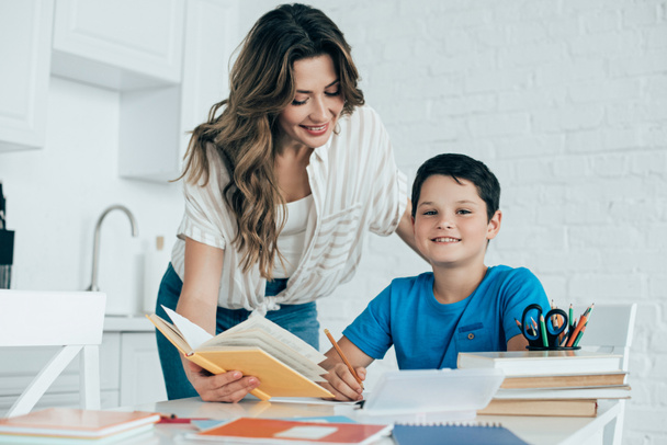 portrait of mother helping smiling son with homework in kitchen at home - Foto, Imagem