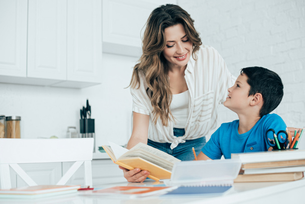 portrait of smiling mother helping son with homework in kitchen at home - Foto, Imagem