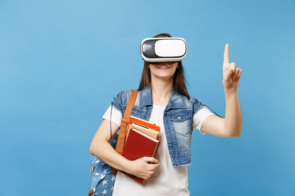 Młoda kobieta studentka w zestaw wirtualnej rzeczywistości trzymać książki touch coś nacisnąć na przycisk, wskazując na pływające wirtualnego ekranu na białym tle na niebieskim tle. Edukacja w szkole university college - Zdjęcie, obraz
