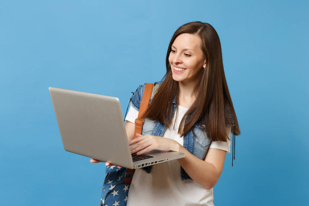 Young beautiful smiling woman student in denim clothes with backpack holding and using working on laptop pc computer isolated on blue background. Education in university. Copy space for advertisement - Photo, image