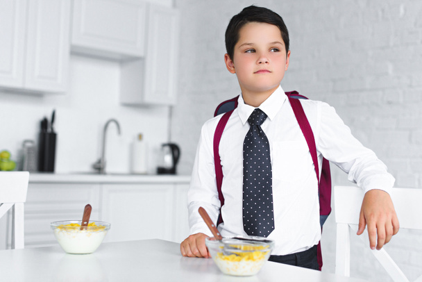 portrait of pensive boy in school uniform with backpack standing at table with breakfast in kitchen, back to school concept - Fotografie, Obrázek