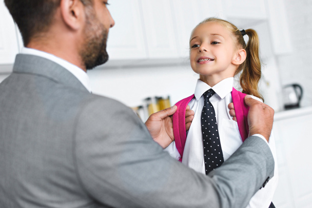 father in suit and little daughter in school uniform with backpack in kitchen at home, back to school concept - Photo, Image