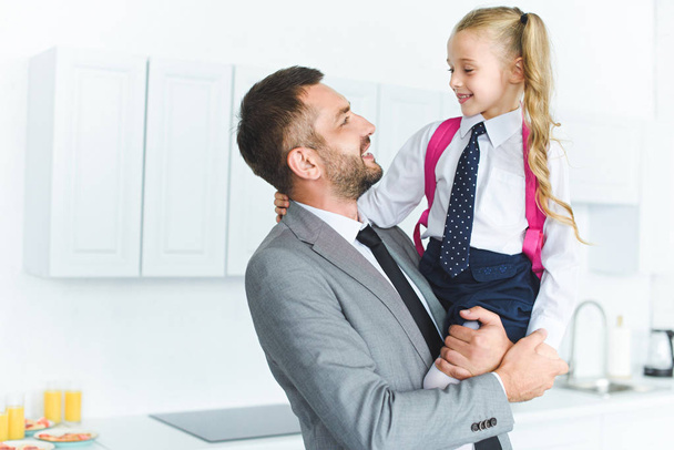 portrait of happy father in suit holding daughter in school uniform with backpack on hands in kitchen, back to school concept - Foto, Bild
