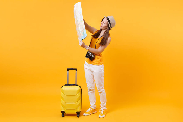 Concentrated tourist woman in summer casual clothes, hat with suitcase looking on city map isolated on yellow background. Female traveling abroad to travel on weekends getaway. Air flight concept - Photo, Image