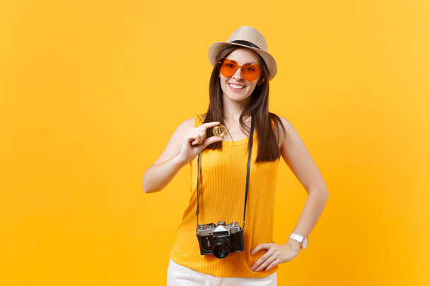 Mulher turística em roupas casuais de verão, chapéu segurando bitcoin, moeda metálica de cor dourada isolada no fundo laranja amarelo. Mulher viajando para o exterior para viajar nos fins de semana de fuga. Conceito de voo aéreo
 - Foto, Imagem