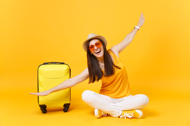 Tourist woman in summer clothes, hat sit at suitcase crossed legs spreading hands as in flight isolated on yellow orange background. Passenger traveling abroad on weekend getaway. Air journey concept - Zdjęcie, obraz