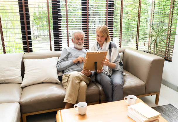 Dos personas mayores disfrutando pasar tiempo juntos mientras están sentados en el sofá y mirando viejas fotografías en el marco de la foto en casa
 - Foto, imagen