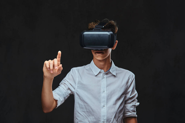 Young student dressed in a white shirt using virtual reality glasses. Isolated on the dark background. - Photo, Image