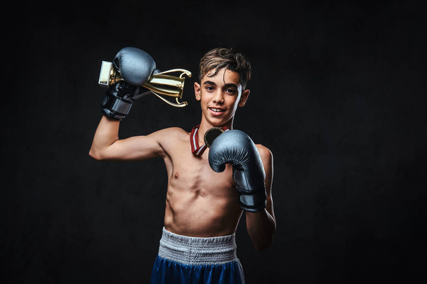 Радісний молодий чемпіон без сорочки з боксу в рукавичках тримає кубок переможця і золоту медаль. Ізольовані на темному тлі
. - Фото, зображення