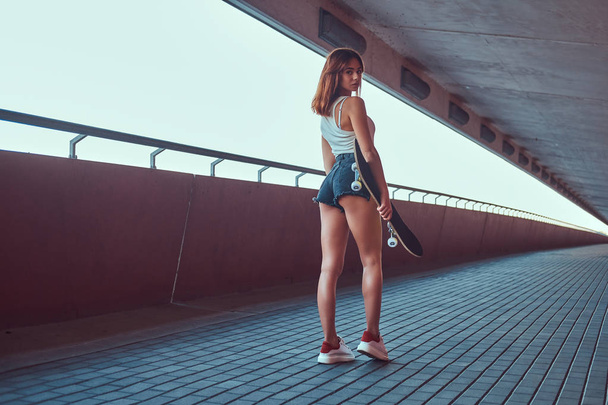 Visão traseira de uma sensual jovem skatista vestida com shorts e camiseta segura um skate e olhando para uma câmera
. - Foto, Imagem