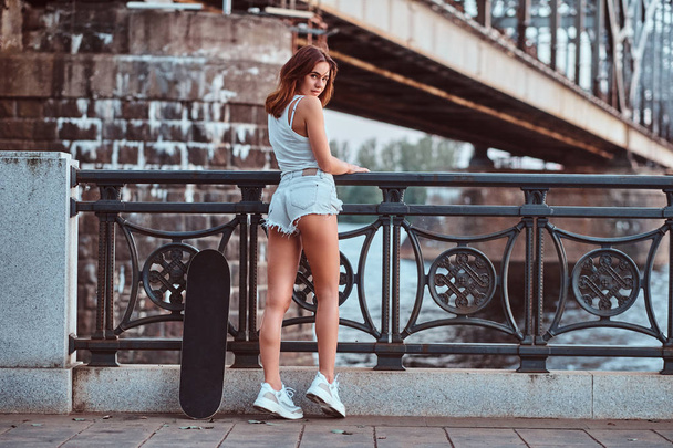 Back view of a sensual skater girl dressed in shorts and t-shirt leaning on the guardrail at a embankment. - Fotografie, Obrázek