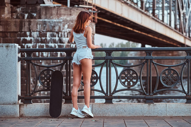 Back view of a sensual skater girl dressed in shorts and t-shirt leaning on the guardrail at a embankment. - Fotó, kép