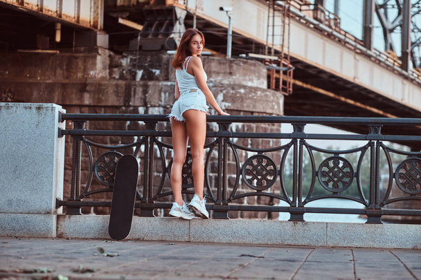 Visão traseira de uma garota patinadora sensual vestida com shorts e camiseta apoiada no corrimão em um aterro
. - Foto, Imagem