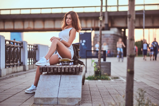Молода дівчина чуттєвого ковзаняра, одягнена в шорти і футболку, сидить на набережній під час красивого заходу сонця
. - Фото, зображення