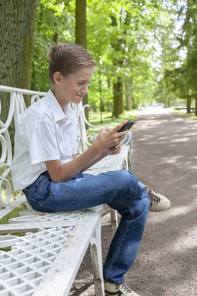 the boy plays in phone in the park in clear summer day - Фото, изображение