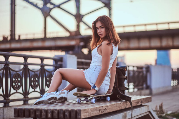 Jovem sensual skater menina vestida com shorts e t-shirt sentado no dique durante belo pôr do sol
. - Foto, Imagem
