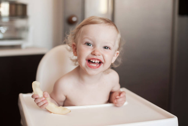 Baby girl eating Honeydew Melon. Funny  laughing child in the kitchen. Cheerful kid in the high chair - Zdjęcie, obraz