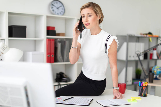 Μια νεαρή ξανθιά κοπέλα κάθεται στο γραφείο του στο γραφείο και μιλάει στο τηλέφωνο. - Φωτογραφία, εικόνα
