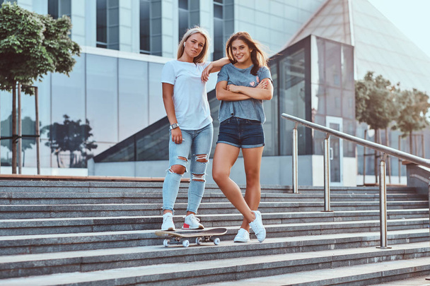 高層ビルの背景にスケート ボードでステップの上に立って 2 人の美しい流行に敏感な女の子. - 写真・画像