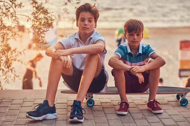 Portrait of two little brothers sitting together on a skateboard against the background of the seacoast at a sunset. - Photo, Image