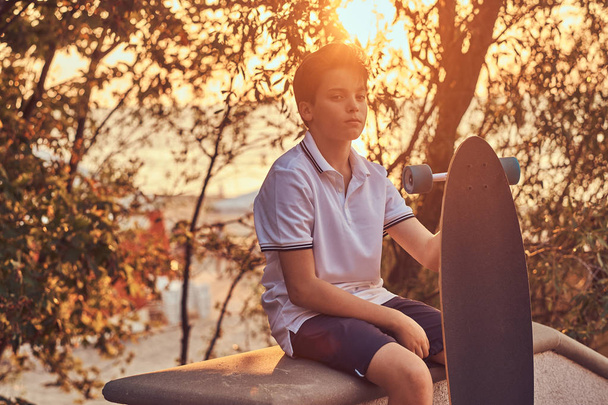 Fiatal deszkás srác öltözött a póló és rövidnadrág ül a kő korláthoz a szabadban egy naplemente.  - Fotó, kép