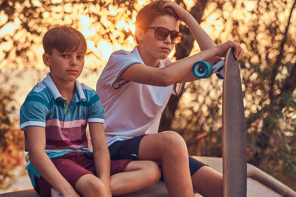Portrét dvou malé bratry se skateboardem sedí na kamenné zábradlí venku na západ slunce. - Fotografie, Obrázek