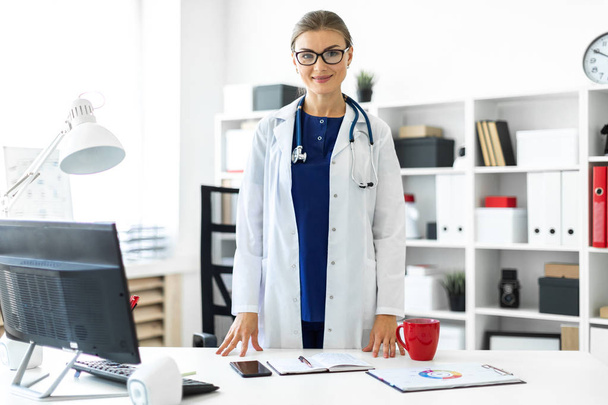 A young girl in a white coat is standing near a table in her office. A stethoscope hangs around her neck. - Photo, Image