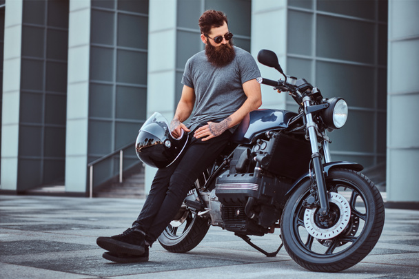 Portrait of a brutal bearded male in a gray t-shirt and black pants holds a helmet sitting on his custom-made retro motorcycle against a skyscraper - Фото, зображення