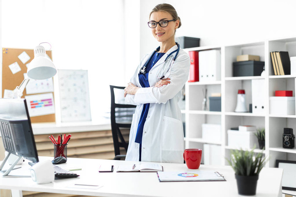 A young girl in a white coat is standing near a table in her office. A stethoscope hangs around her neck. - Photo, Image