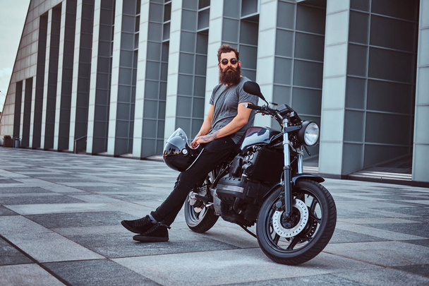 Brutal bearded male in a gray t-shirt and black pants holds a helmet sitting on his custom-made retro motorcycle against a skyscraper. - Photo, Image