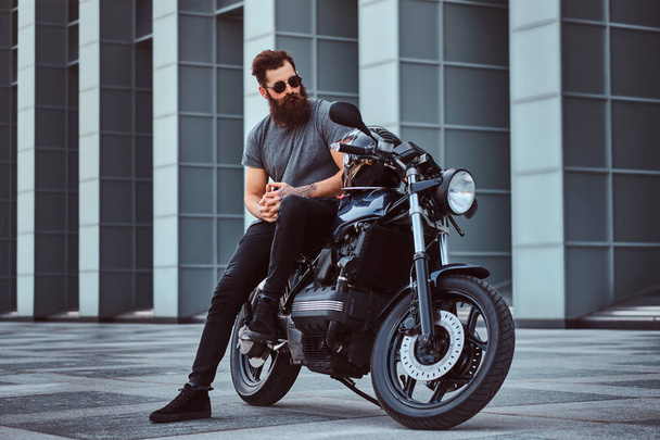 Brutal bearded male in sunglasses dressed in a gray t-shirt and black pants sitting on his custom-made retro motorcycle against skyscraper. - Photo, Image