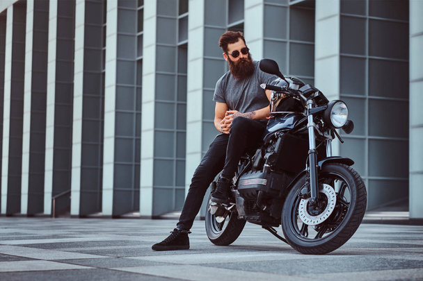 Жорстокий бородатий чоловік в сонцезахисних окулярах, одягнений у сіру футболку і чорні штани, сидячи на своєму спеціальному ретро мотоциклі проти хмарочоса
. - Фото, зображення