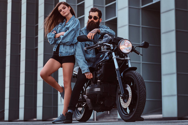 Atraente casal hipster barbudo macho brutal em óculos de sol e jeans jaqueta sentado em uma motocicleta retro e sua jovem menina sensual de pé perto, posando contra arranha-céus
. - Foto, Imagem