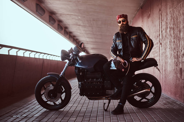 Retrato de un elegante hombre barbudo vestido con una chaqueta de cuero negro y bandana con gafas de sol sentado en su motocicleta retro hecha a medida, mirando a una cámara
. - Foto, Imagen