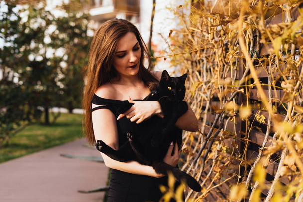 charming long-haired beautiful woman in black dress holds a black cat on her hands, walks in the garden in the evening - Foto, immagini