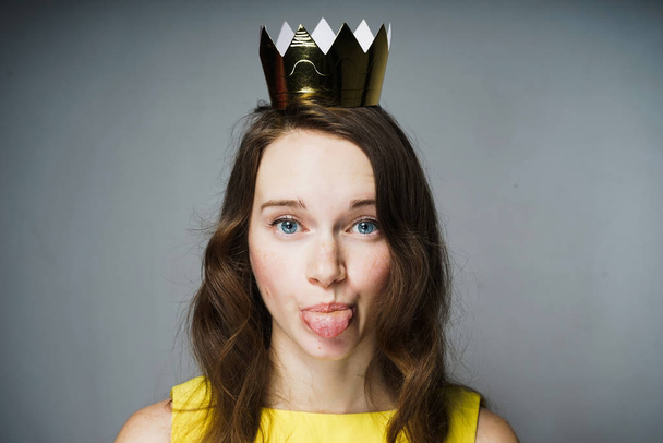 funny blue-eyed girl in a yellow dress shows a tongue, a golden crown on her head - Photo, Image