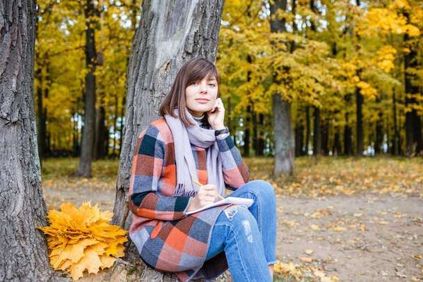 Portrait of young fashion woman outdoor. Writes in a notebook. University Park, study concept. Brunette woman in autumn park with fashionable plaid coat and scarf. - Foto, imagen