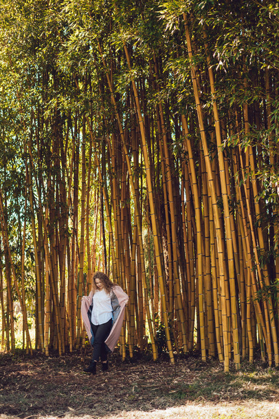 stylish young woman in white shirt and pink coat posing in the park next to bamboo trees, enjoying the warmth - Foto, afbeelding