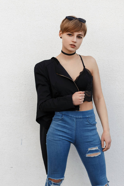 Bella giovane donna modello hipster in biancheria intima nera sexy pizzo, jeans e giacca nera elegante posa vicino al muro bianco
 - Foto, immagini