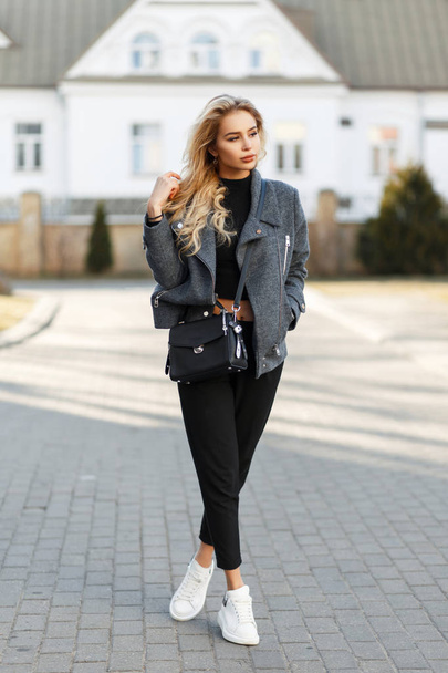 Mooie stijlvolle meisje met een tas in een mode grijze vacht, zwarte broek en witte sneakers in de straat op een zonnige dag - Foto, afbeelding