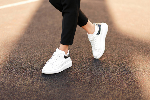 Κομψά μοντέρνα γυναικεία αθλητικά στο δρόμο. Γυναικεία πόδια με λευκά παπούτσια στην άσφαλτο σε μια ηλιόλουστη ημέρα - Φωτογραφία, εικόνα