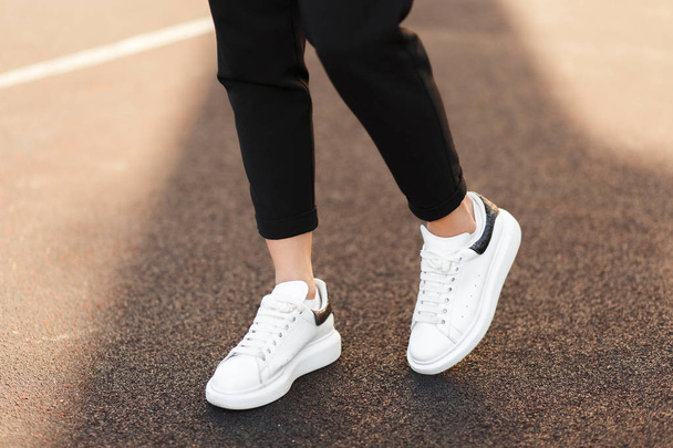 Όμορφα γυναικεία πόδια με λευκά αθλητικά παπούτσια στην άσφαλτο σε μια ηλιόλουστη ημέρα - Φωτογραφία, εικόνα