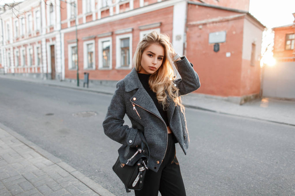 Fashionable beautiful young woman in a stylish gray jacket and black pants with a bag in the city at sunset - Zdjęcie, obraz