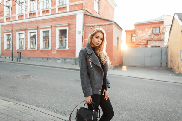 Досить молода модель дівчина зі стильною сумочкою в модному сірому пальто ходить на заході сонця в місті
 - Фото, зображення