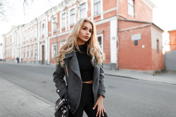 Beautiful young woman model with a bag in a stylish gray jacket walking outdoors in the city - Photo, Image