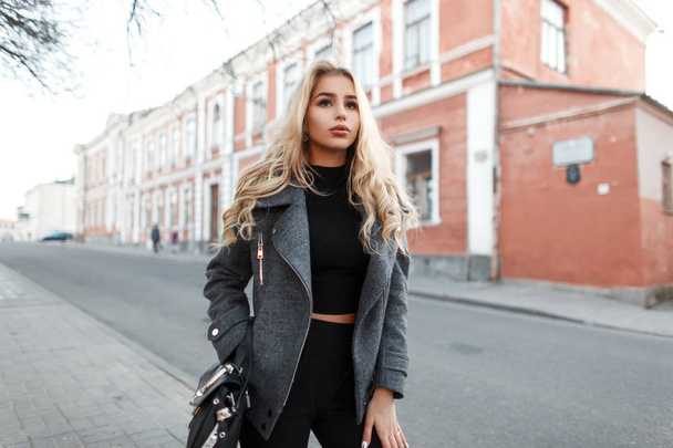 Mulher jovem elegante na moda com uma bolsa preta em uma jaqueta de moda cinza caminha na cidade
  - Foto, Imagem